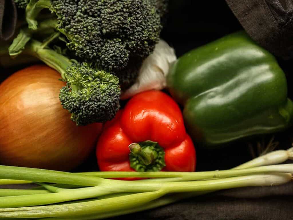 A collection of fresh vegetables: a bunch of broccoli, a green bell pepper, a red bell pepper, an onion, and green onions. They are arranged together on a dark background.